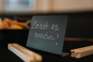 Ein schwarzes Schild mit der handgeschriebenen Aufschrift "Lasst es krachen!" steht auf einem Tisch neben zwei Holzstäbchen. Im Hintergrund ist unscharf eine Schale mit Obst zu sehen.