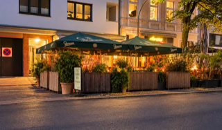 Straßencafé mit grünen Sonnenschirmen und Holzbarrikaden, umgeben von Pflanzen, bei Abendbeleuchtung.